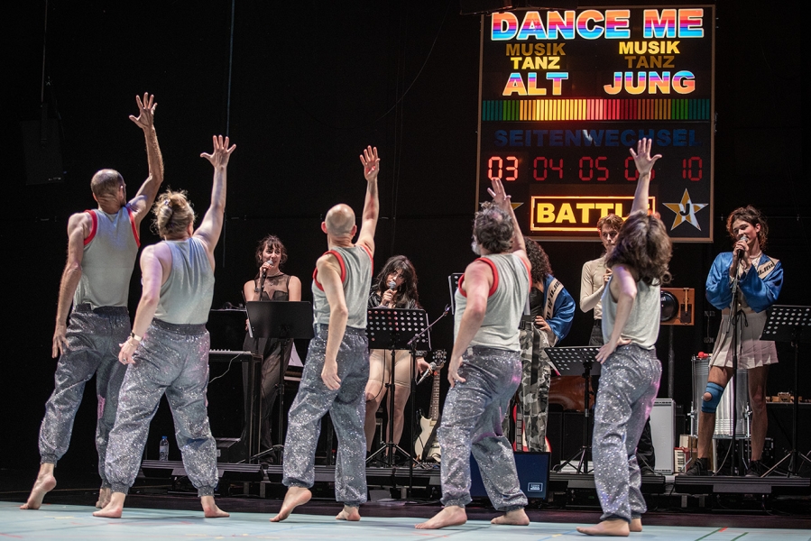 Photo from the She She Pop Show Dance Me featuring 5 old and 5 young performers. We see 5 older people in sparkly trousers and silver grey vests with their right hands in the air in front of 5 younger performers behind microphone stands. The background is the illuminated scoreboard from the show, designed by Jan Brokof